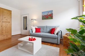 A living room with a grey sofa, red and white pillows, a white coffee table, a wooden wardrobe, and a painting of flowers on the wall.
