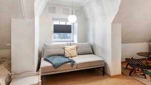 A loft-style bedroom with a built-in sofa, a small dining table, and a window with blinds.