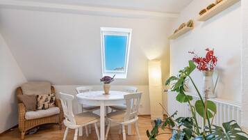 A dining area with a round table, white chairs, a wicker chair, a window, and a shelf with decorative items.
