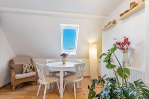 Un coin repas comprenant une table ronde, des chaises blanches, un fauteuil en osier, une fenêtre et une étagère ornée d’objets décoratifs.