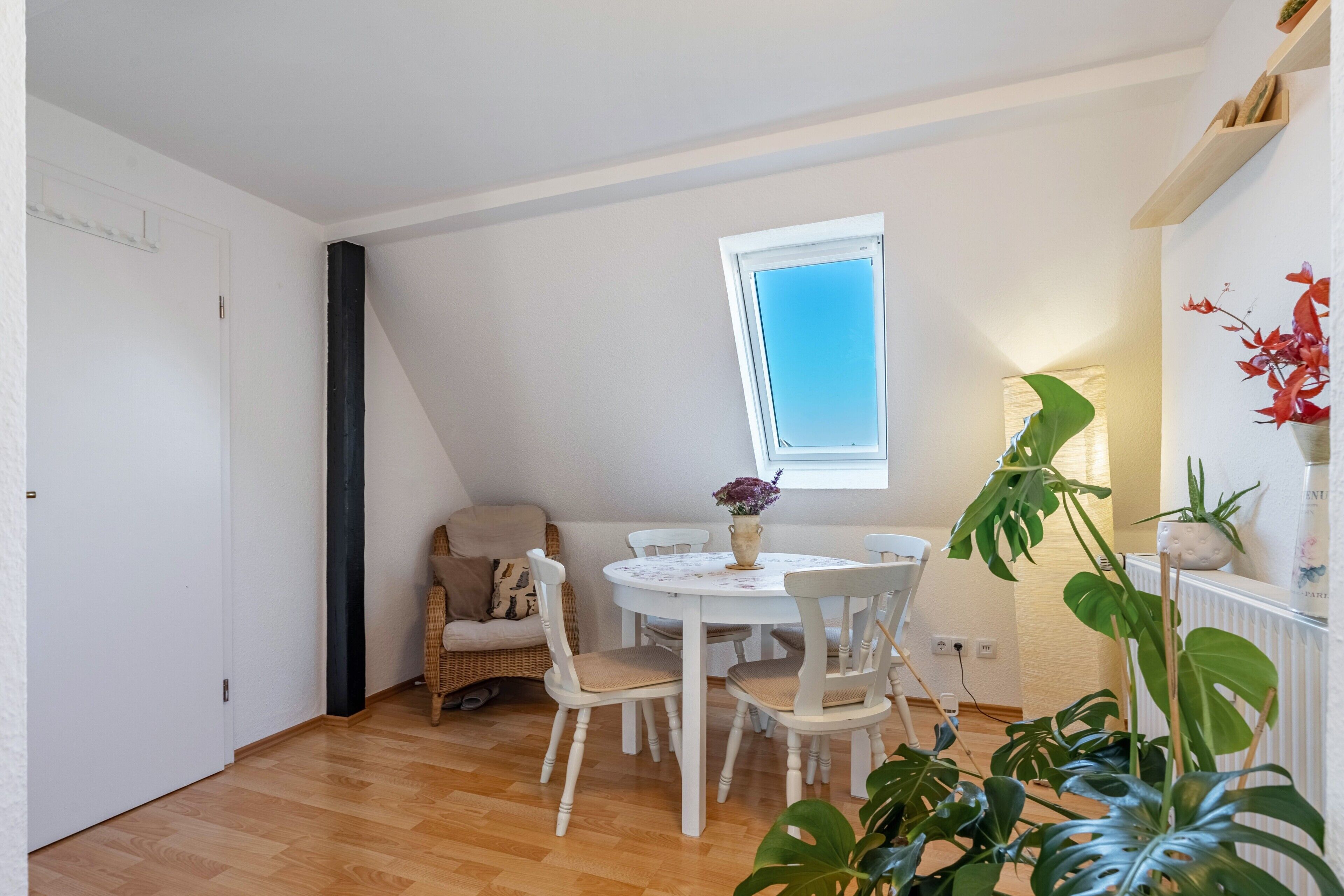 Ein Essbereich mit rundem Tisch, weißen Stühlen, einer Rattan-Couch, einem Fenster und Topfpflanzen.
