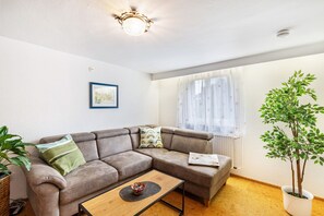 Un salon comprenant un canapé d’angle, une table basse, une plante en pot, un tableau encadré accroché au mur et une fenêtre ornée de rideaux légers.