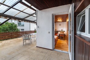Eine überdachte Terrasse mit einer hölzernen Bank, einer weißen Wand und Blick in einen Raum mit Bett und Tisch.