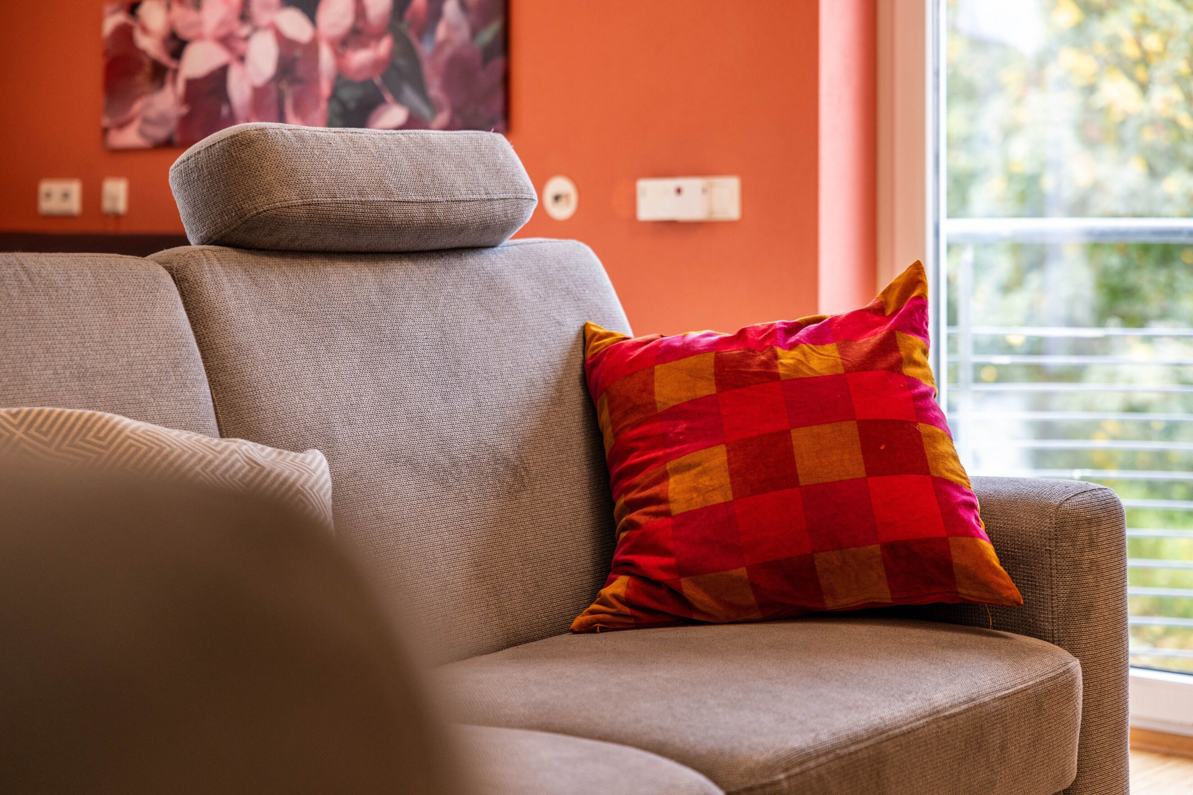 A sofa with a checkered pillow, a wall outlet, and a window with a view.