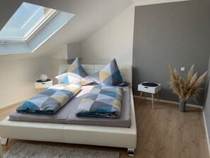 Une chambre à coucher moderne avec une lucarne, un lit recouvert d’une literie à motifs, une table de chevet, une petite table d’appoint et un vase contenant des plumes d’herbe des pampas.