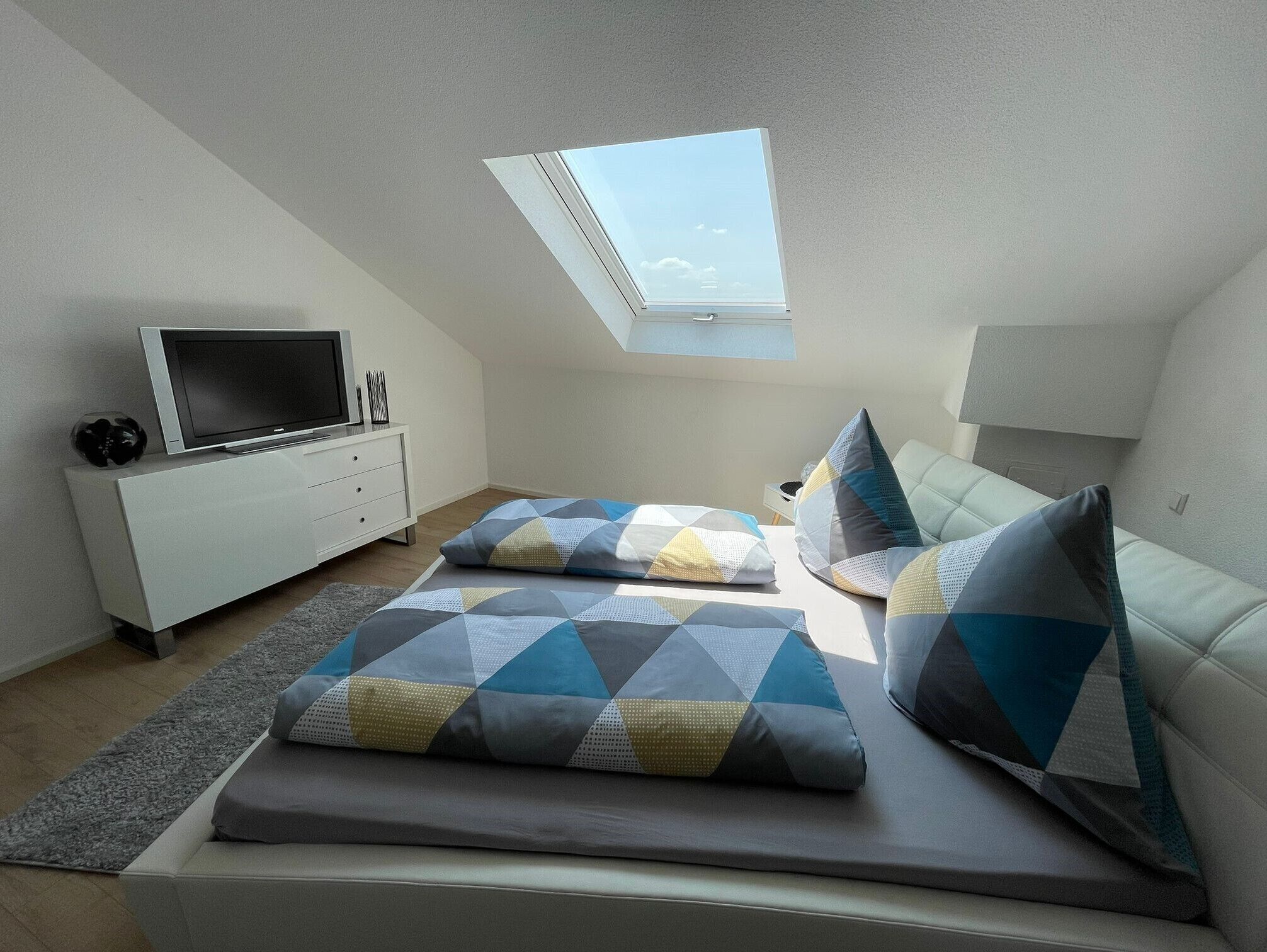 Une chambre avec un puits de lumière, un téléviseur posé sur un meuble blanc, un lit agrémenté d’oreillers à motifs et un canapé de couleur claire.