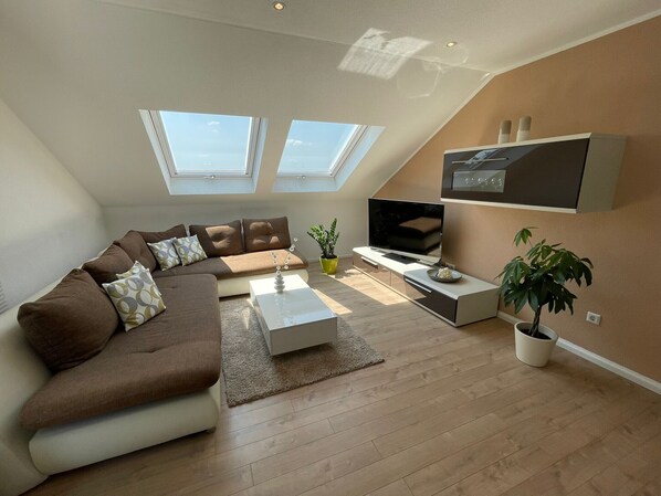 Un salon moderne avec un canapé d’angle, un téléviseur à écran plat, une table basse et une plante en pot.
