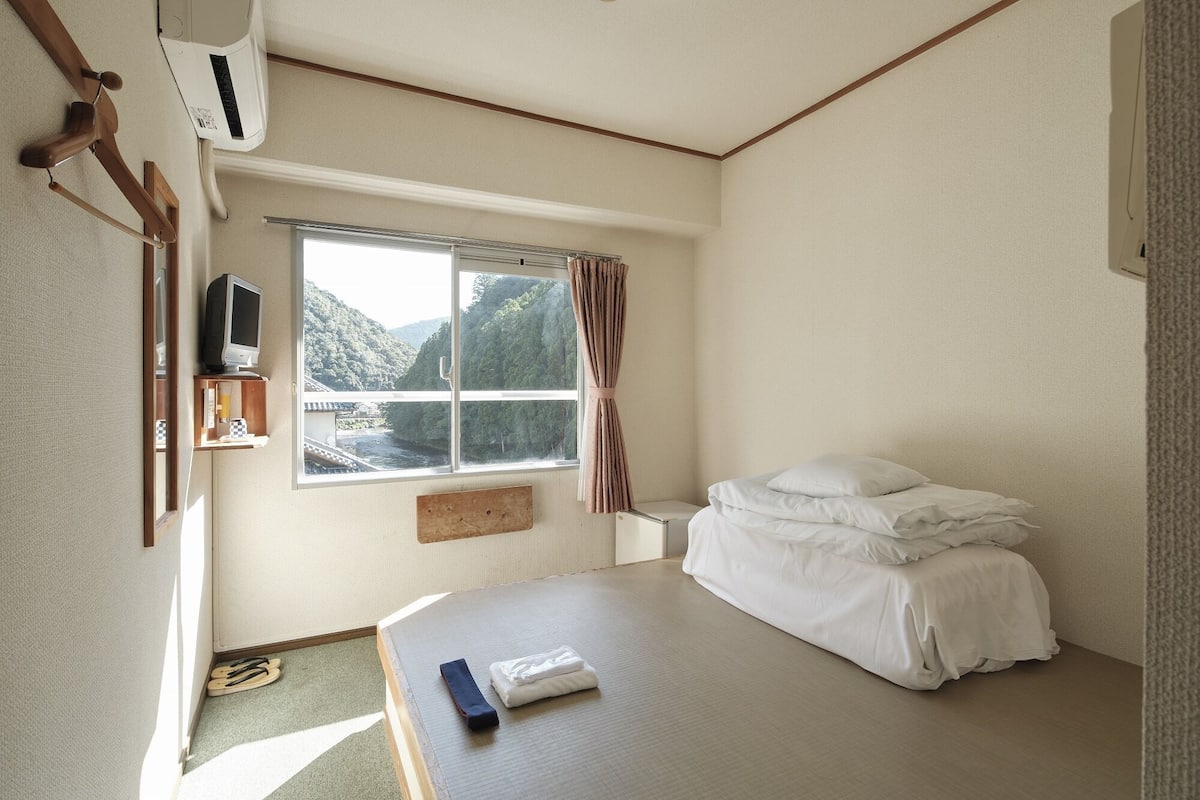 A bedroom with a bed, a window with a view of a river and hills, a TV, and a small shelf with items.