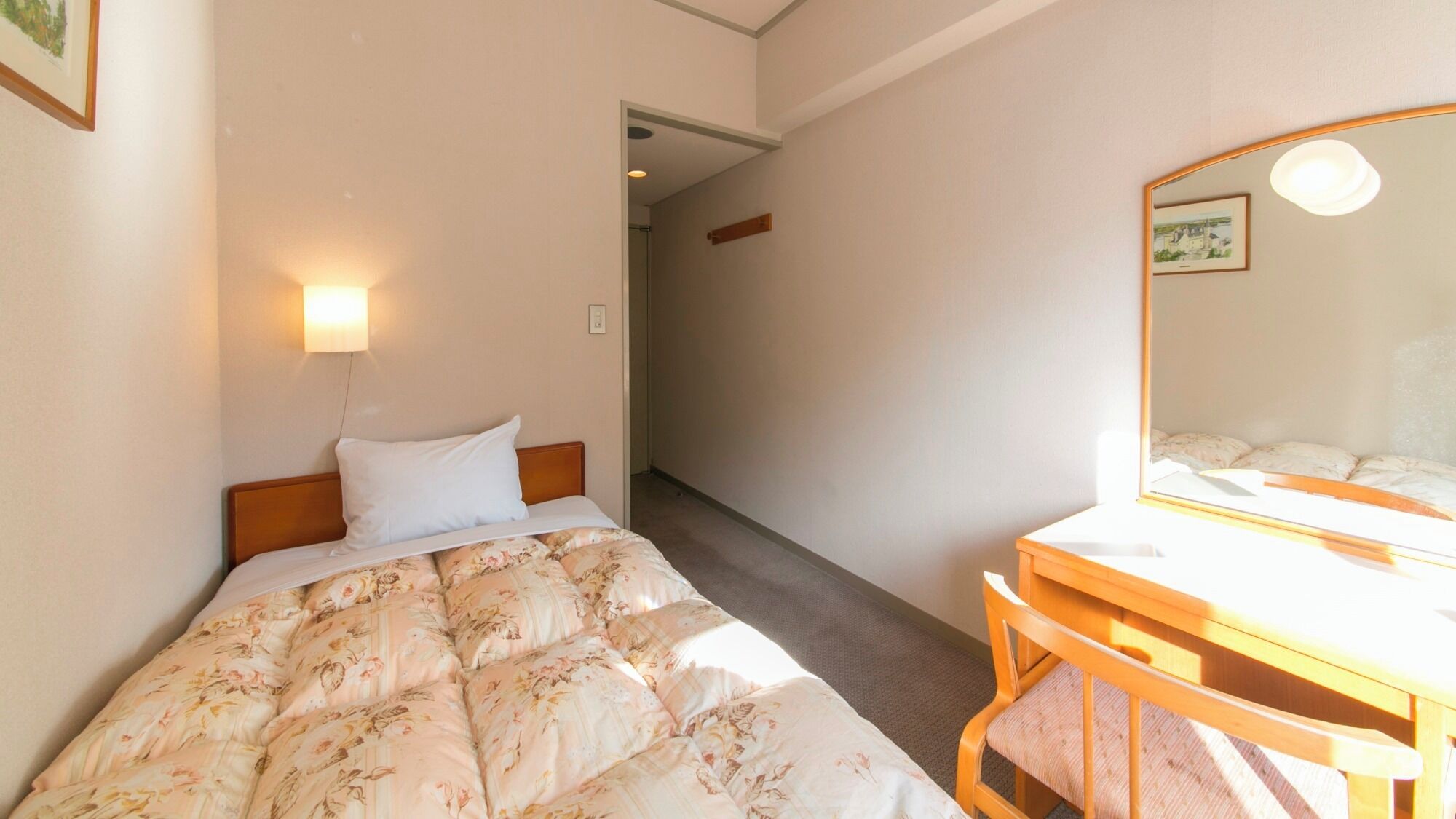 A hotel room with a bed, a bedside table, a chair, a mirror, and a hallway leading to another room.