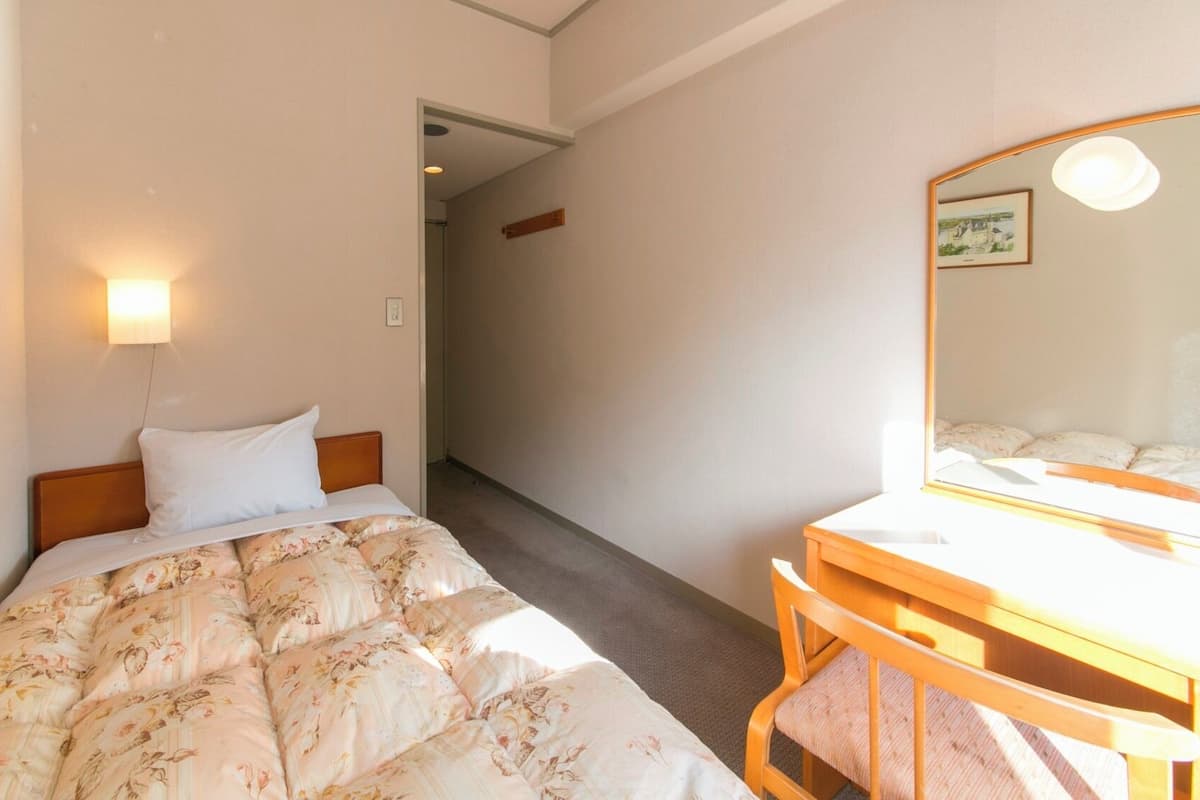 A hotel room with a bed, a bedside table, a chair, a mirror, and a hallway leading to another room.