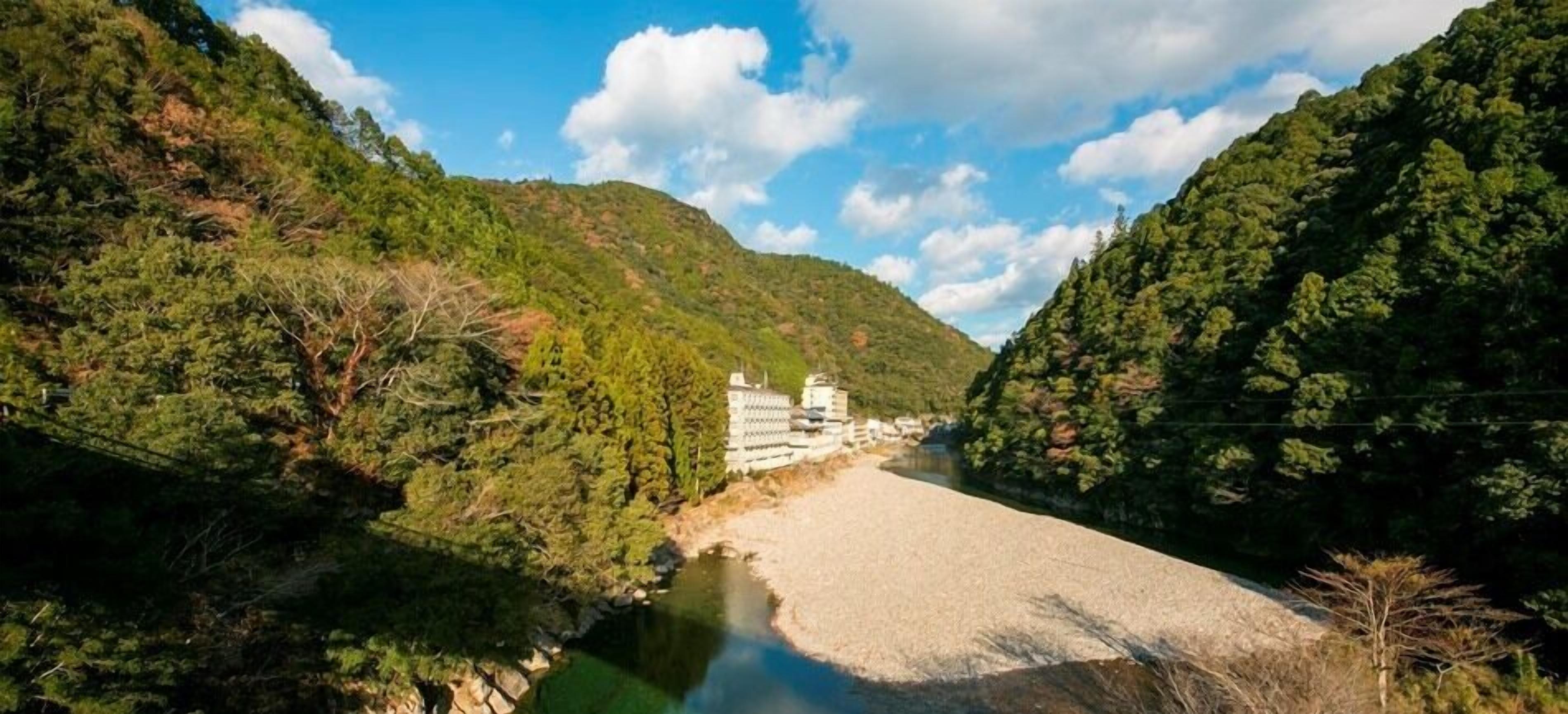 A river with a bridge, surrounded by dense green forest and hills.