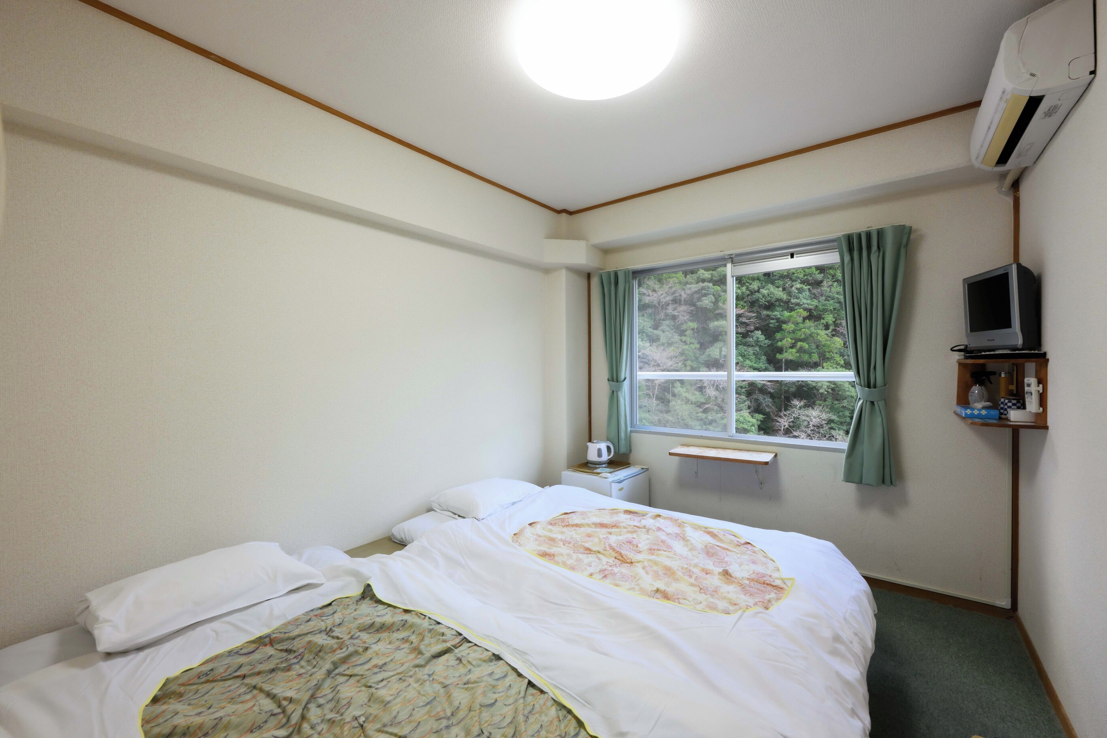 A small, simple room with a bed, a window with curtains, a small table with a kettle, and a television on a stand.