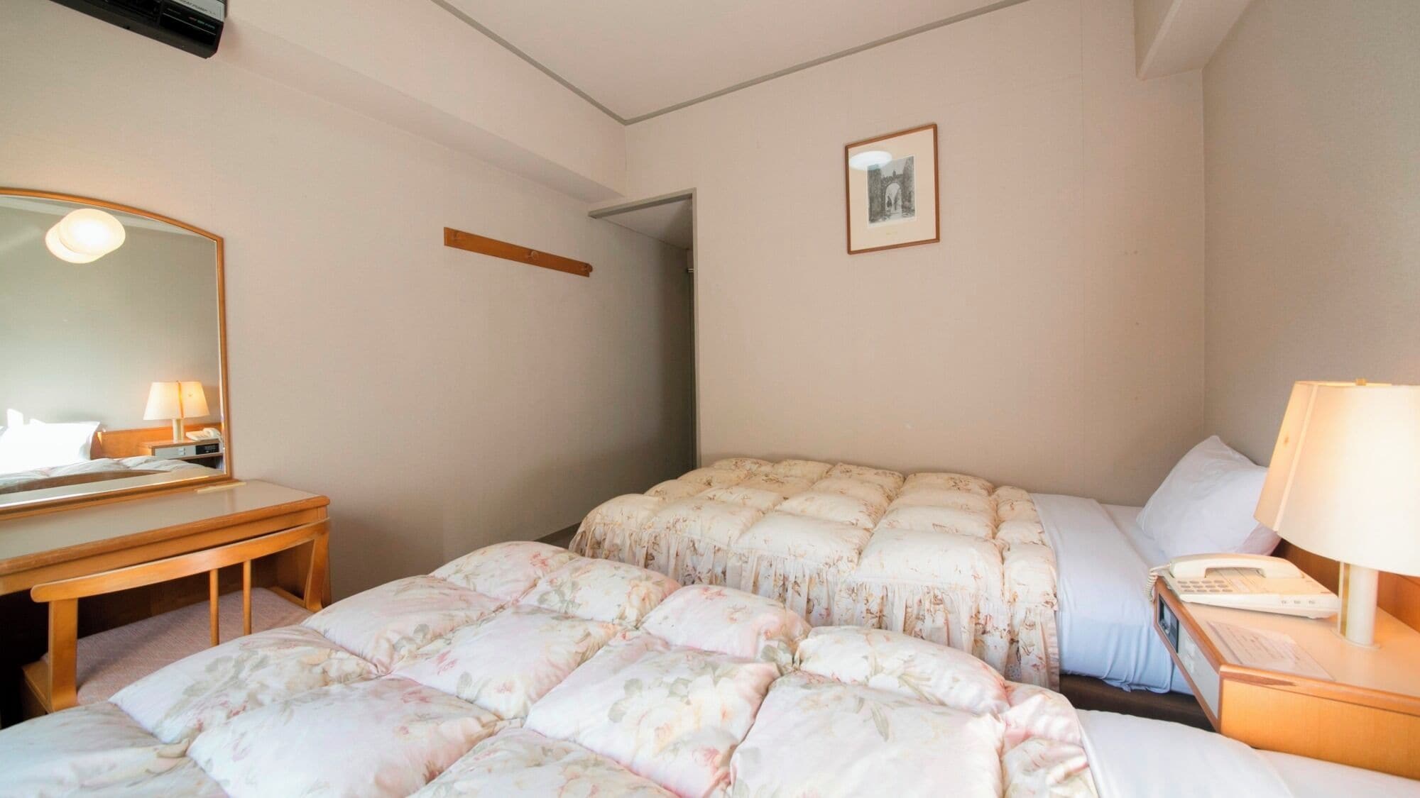 A hotel room with a bed, a desk, a chair, a bedside table with a lamp, and a framed picture on the wall.