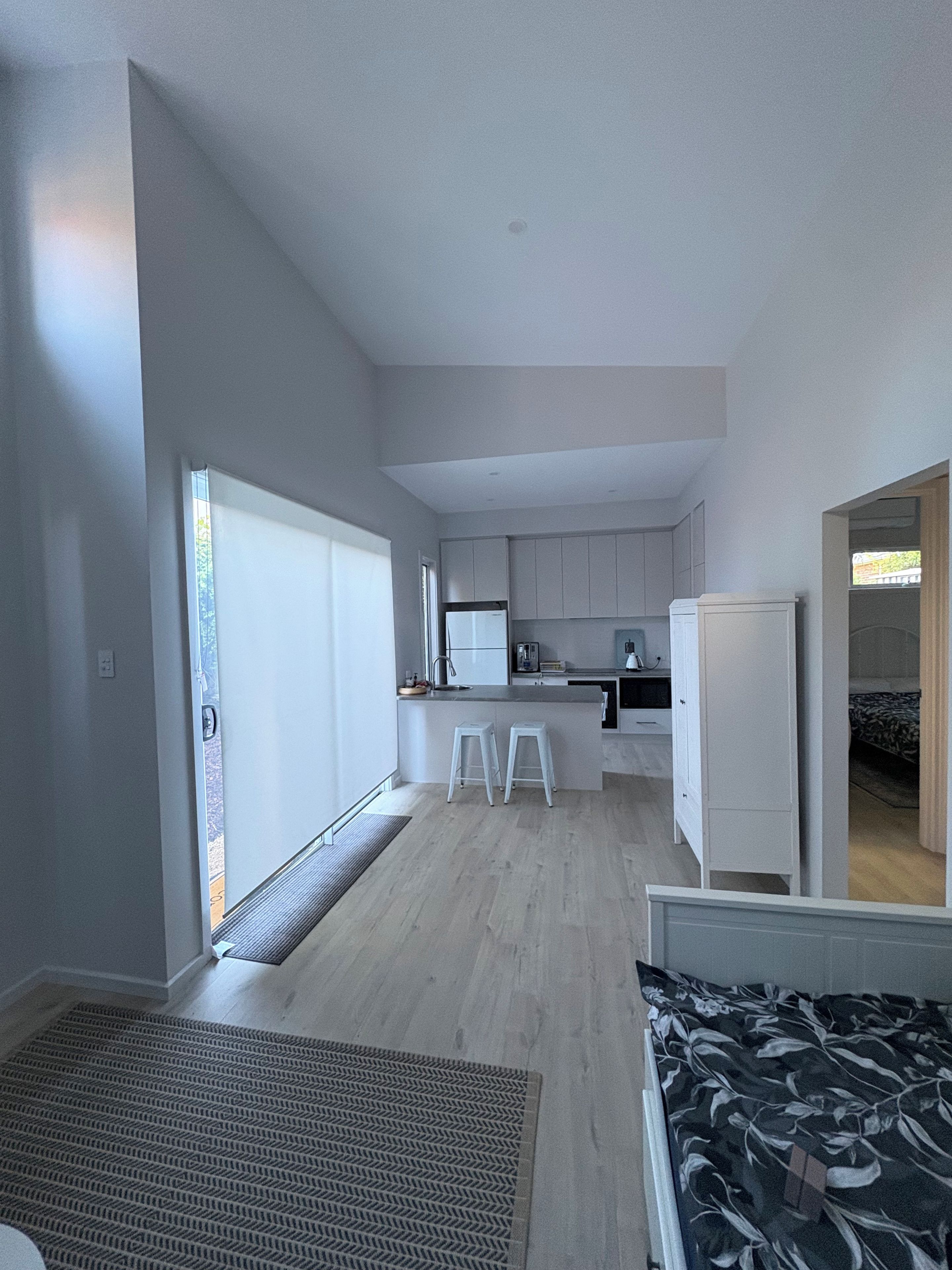 A modern, minimalist bedroom with a large bed, a small sitting area, and a kitchenette in the background.
