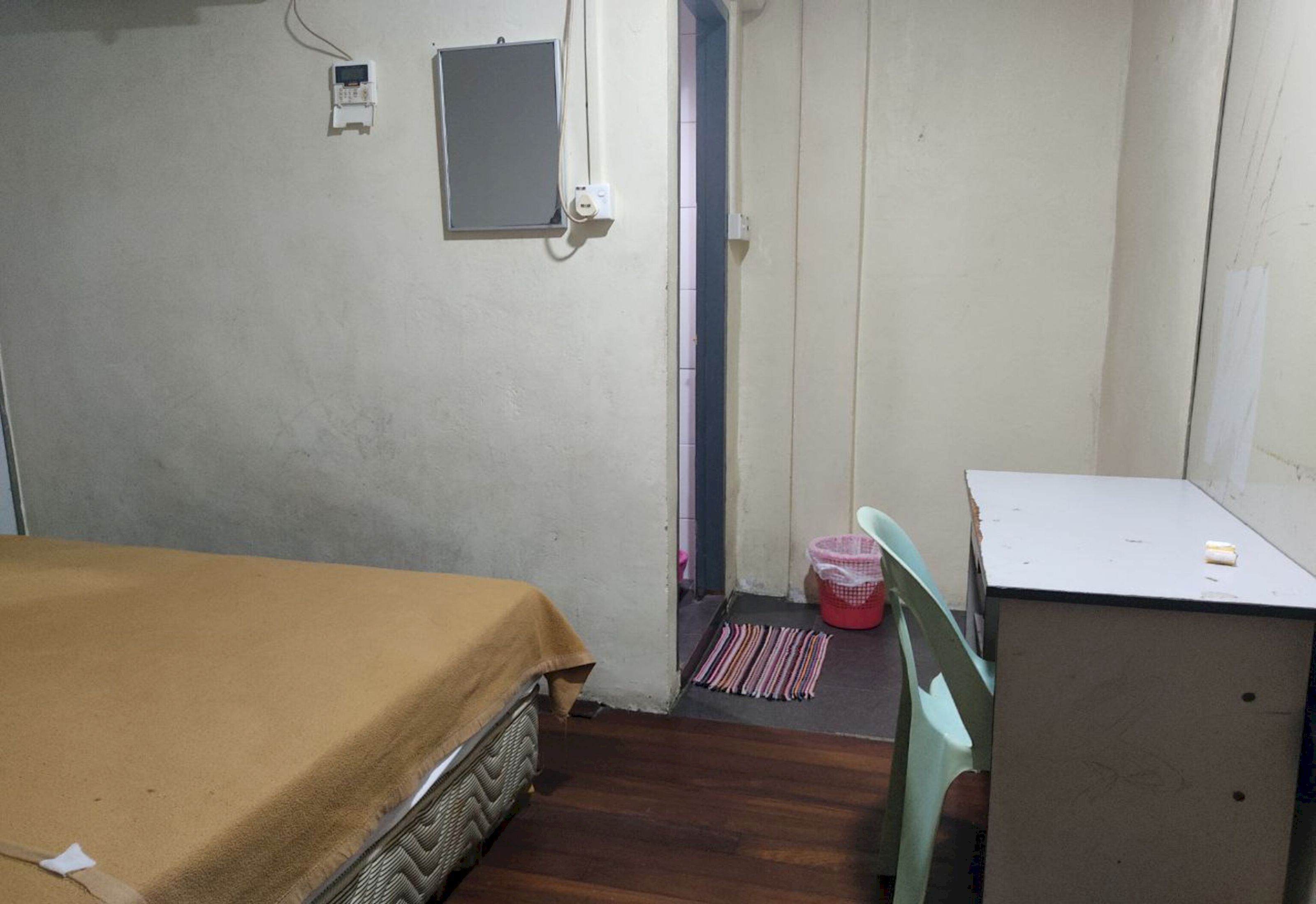 A small, sparsely furnished room with a bed, a desk, and a chair. There is a door leading to another room, a small electrical outlet, and a trash bin.