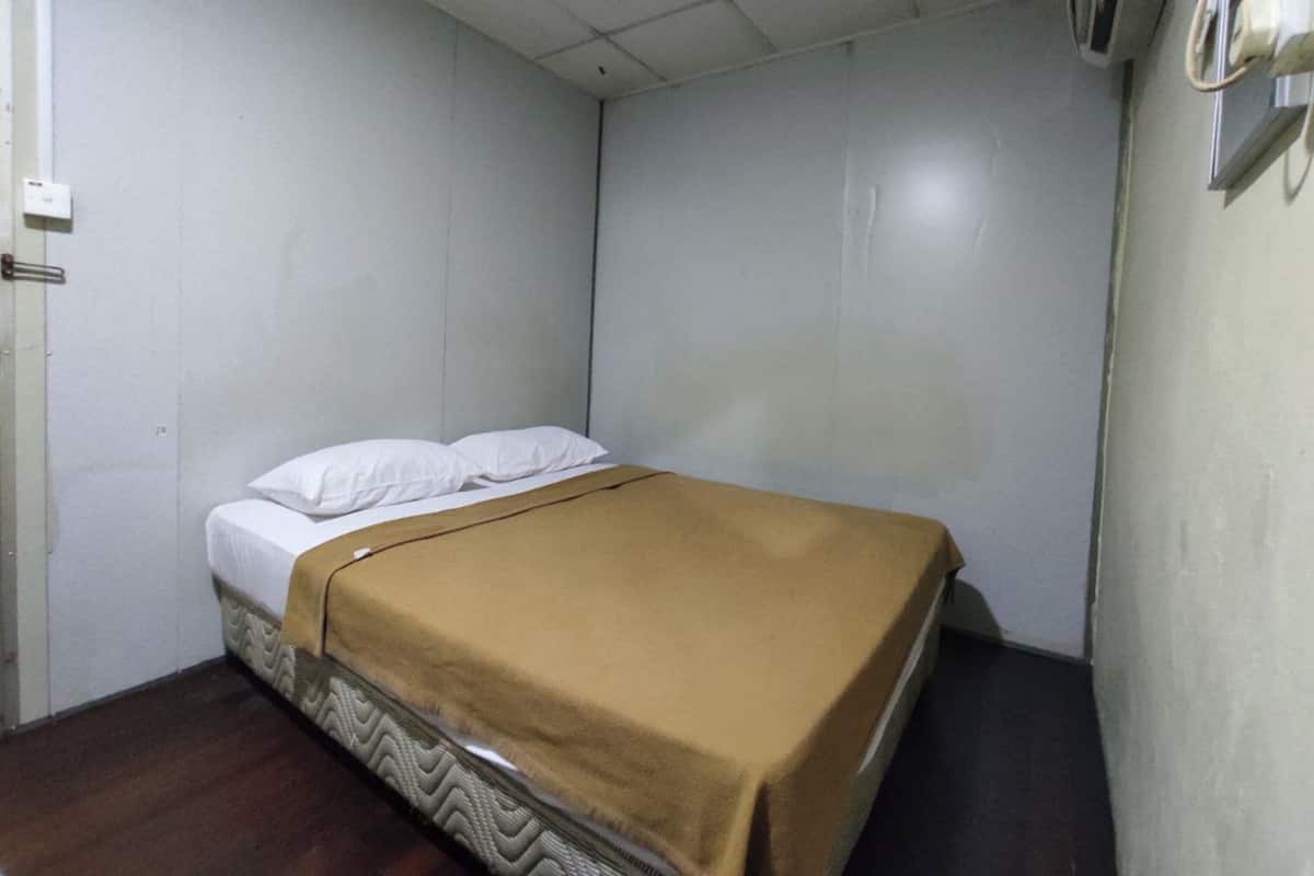 A small, single-bed room with a plain white wall, a brown bedspread, and a visible air conditioning unit.