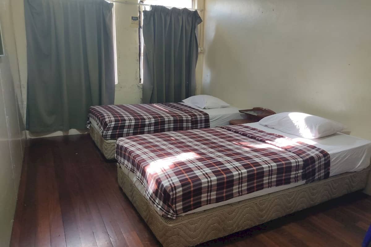 A room with two beds, each with a plaid bedspread and white pillows. There is a window with curtains, and the room has wooden flooring.