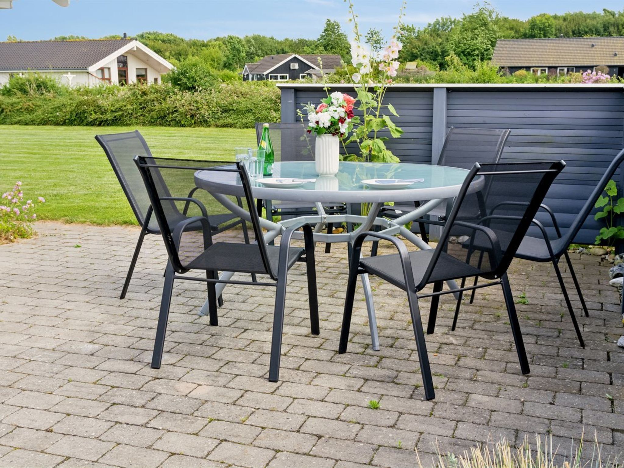 A patio with a round glass table and four chairs, surrounded by a brick floor and a garden.