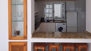 A kitchen with a built-in washing machine, a sink, and a window with blinds. There are wooden stools and a glass cabinet with wine glasses. - La Marea Casa en Arrieta, Tranquila. Cerca de Restaurantes y Playas de Arena (CN)