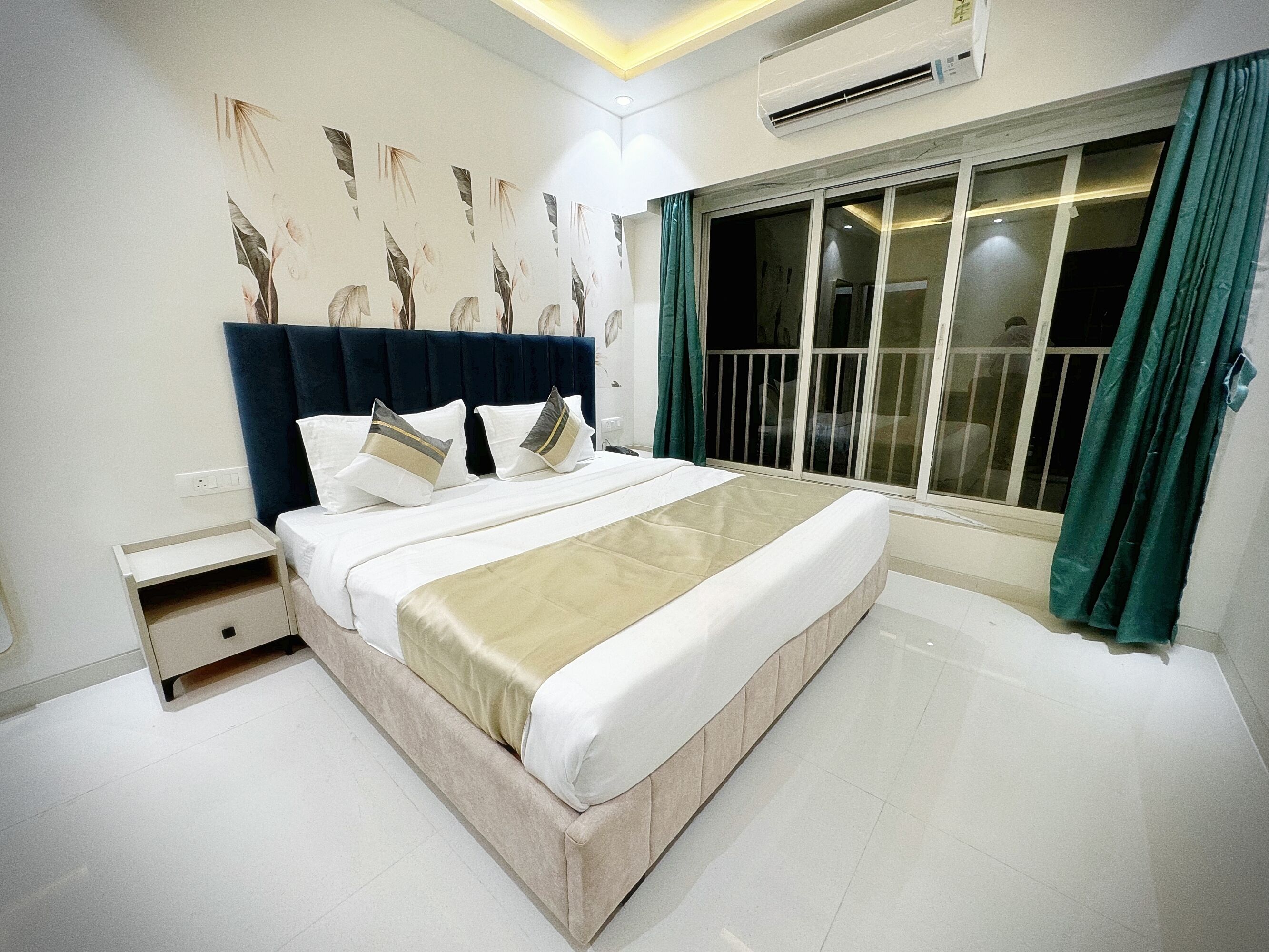 A modern bedroom with a large bed, a nightstand, a wall-mounted air conditioner, and a view through a sliding door.