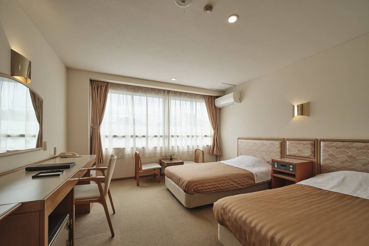 A hotel room with a desk, two beds, a chair, and a window with curtains.