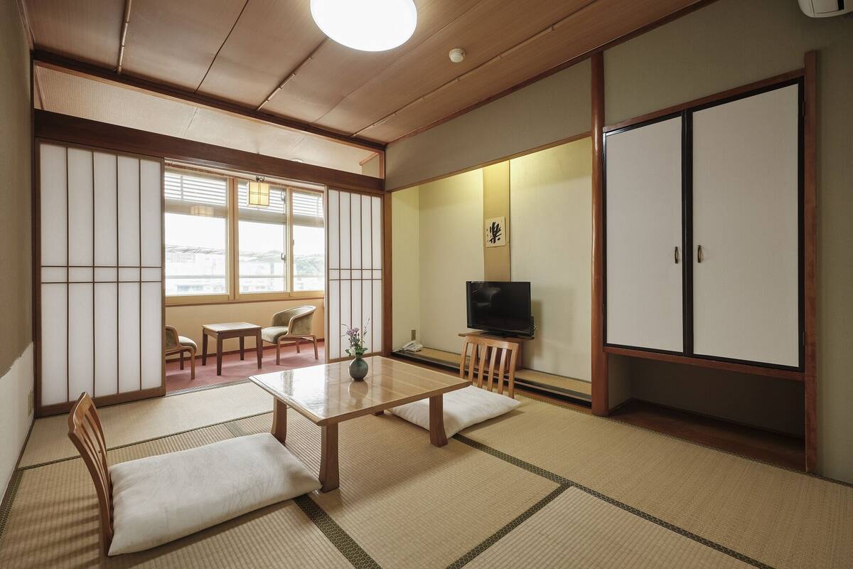 A traditional Japanese room with tatami flooring, a low table with cushions, a television on a small stand, and sliding doors.