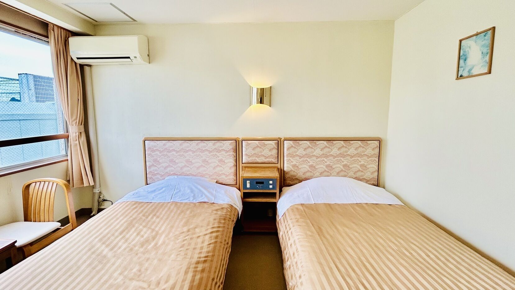 A hotel room with two beds, a small table, a chair, an air conditioning unit, and a wall-mounted light fixture.