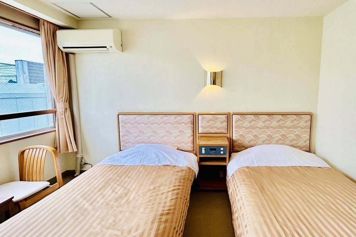 A hotel room with two beds, a small table, a chair, an air conditioning unit, and a wall-mounted light fixture.
