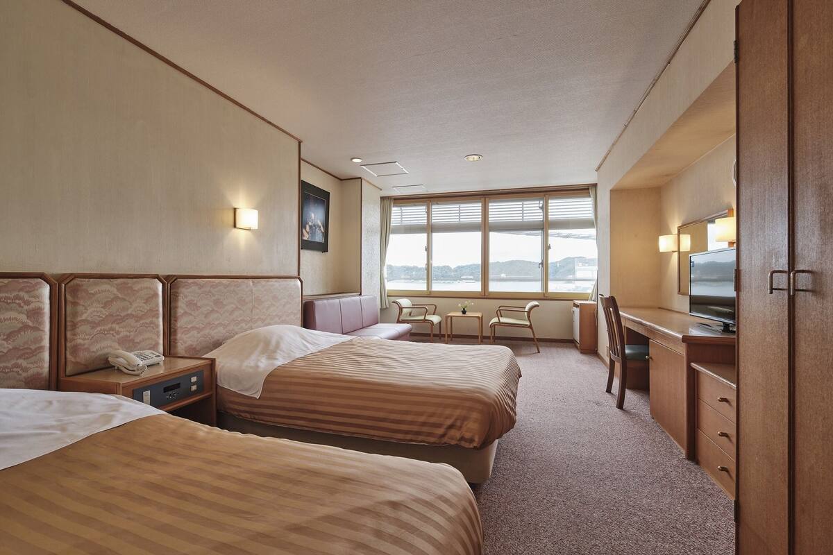 A hotel room with two beds, a desk, a chair, a TV, and a large window with a view.
