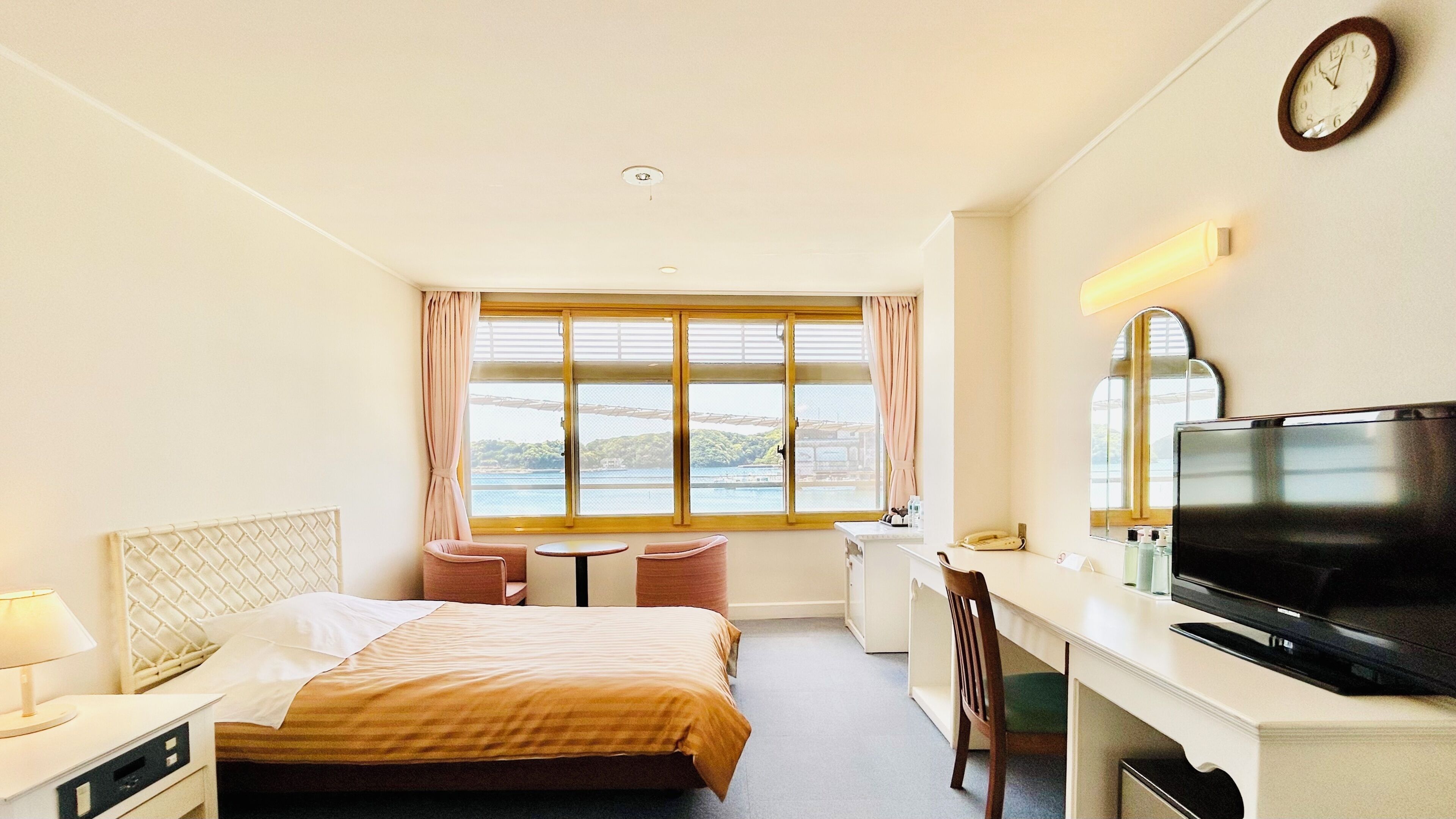 A hotel room with a bed, a desk with a TV, a chair, a window with a view, and a clock on the wall.