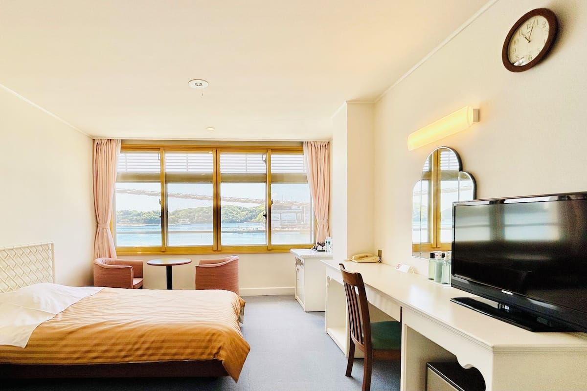A hotel room with a bed, a desk with a TV, a chair, a window with a view, and a clock on the wall.