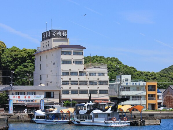 En byggnad med flera våningar, en skylt på japanska, en båt som ligger förtöjd framför och ett skogbeklätt kullområde i bakgrunden.