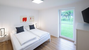 Ein modernes Schlafzimmer mit einem großen Bett, einem Nachttisch, einem an der Wand befestigten Fernseher und Blick auf eine grüne Außenanlage durch eine Schiebetür.