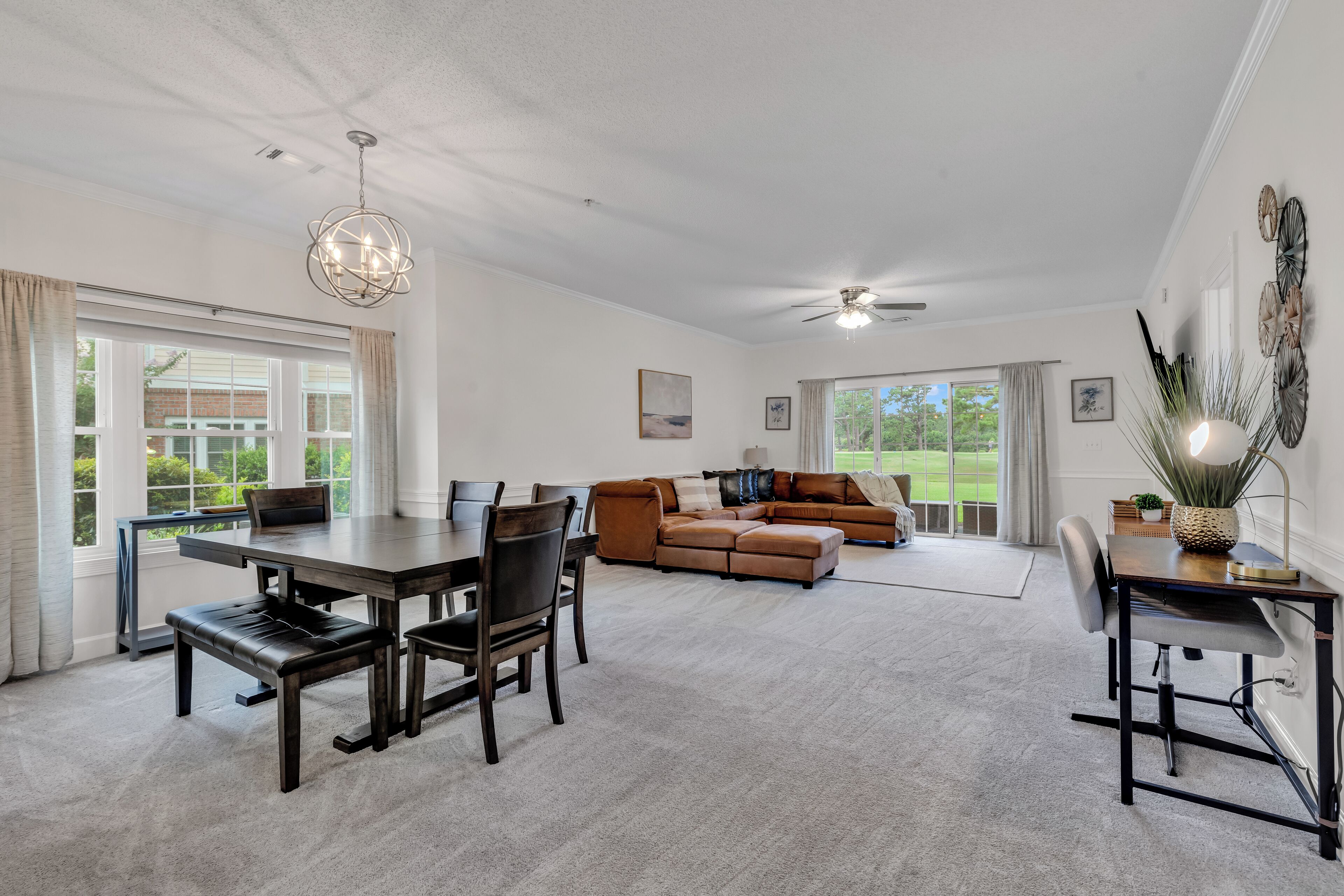 A spacious living room with a dining area, a ceiling fan, and large windows offering a view of the outdoors.