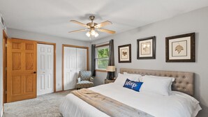 A bedroom with a large bed, a ceiling fan, framed pictures on the wall, and a window with curtains. - Charming Family Home in Burnsville (Burnsville)