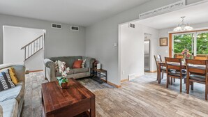 A spacious living area with a grey sofa, a wooden coffee table, and a dining table set with chairs. There is a staircase leading to another level, and a window providing a view of greenery outside. - Charming Family Home in Burnsville (Burnsville)