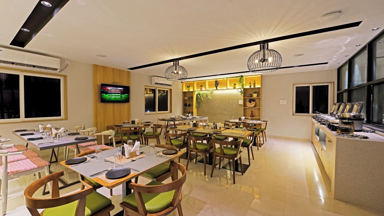 A restaurant with multiple dining tables and chairs, a television mounted on the wall, and a kitchen area visible through an open partition.