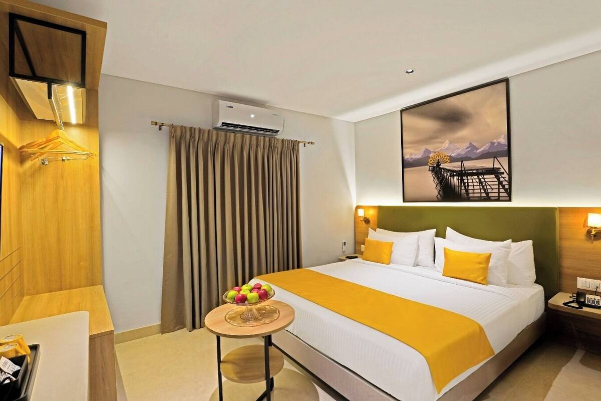 A hotel room with a bed, a TV, a small table with fruit, and a painting on the wall.
