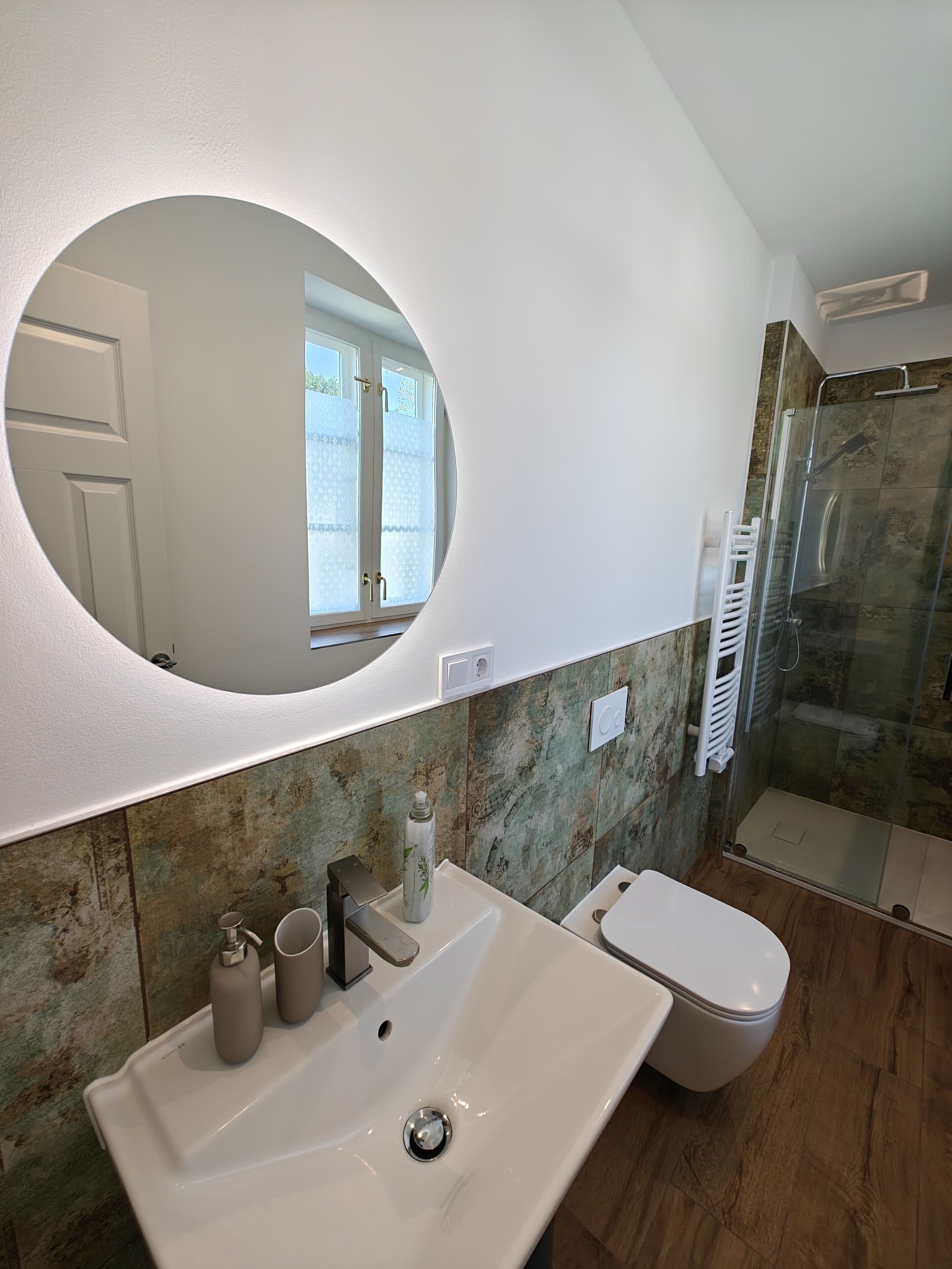Une salle de bain avec un grand miroir ovale, un lavabo blanc, des toilettes et une cabine de douche en verre.