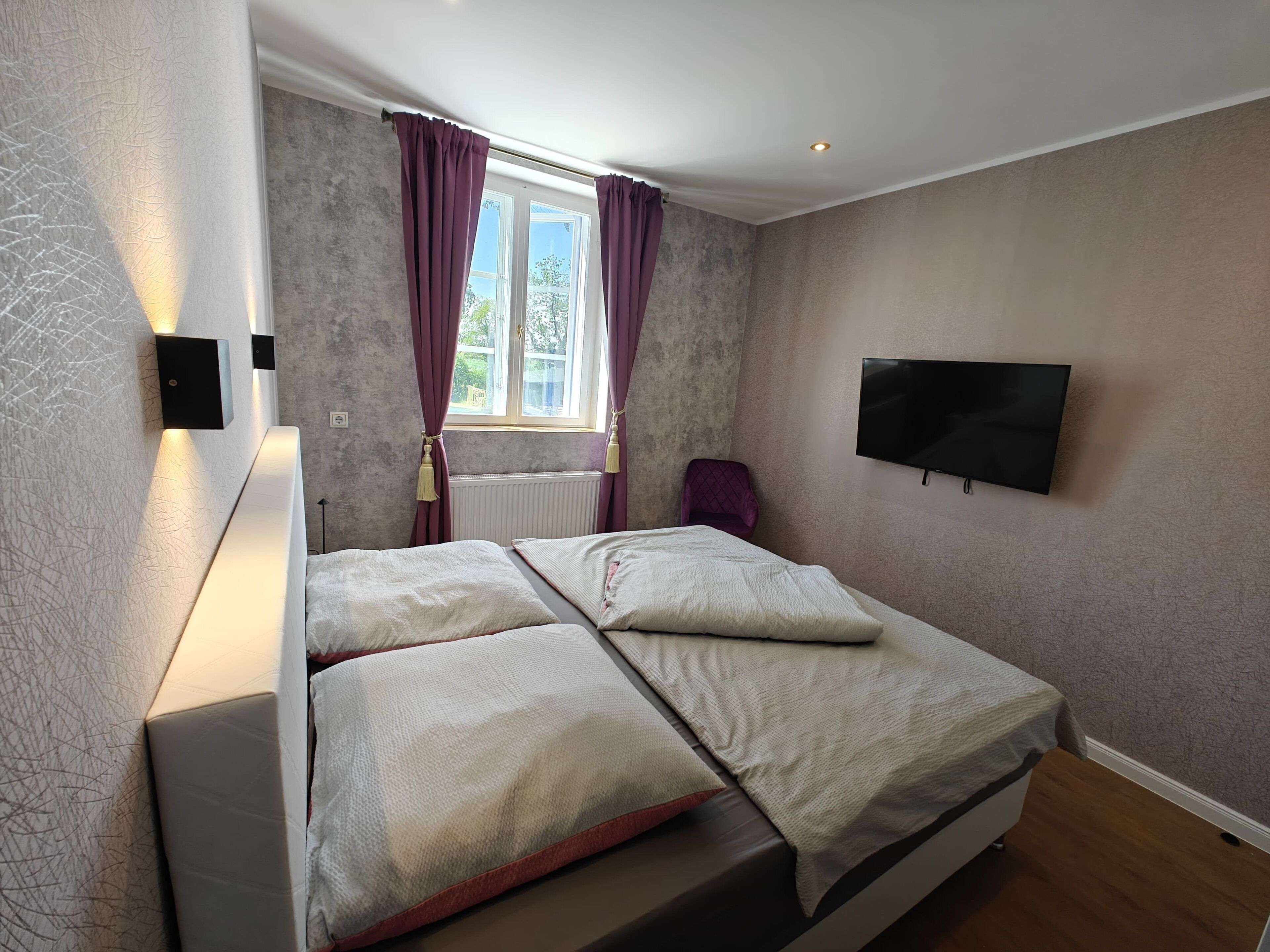A bedroom with a bed, a TV, a window with curtains, and a radiator.