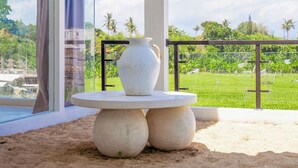 A modern outdoor seating area with a white table and vase, surrounded by sand, overlooking a green landscape and a clear sky.