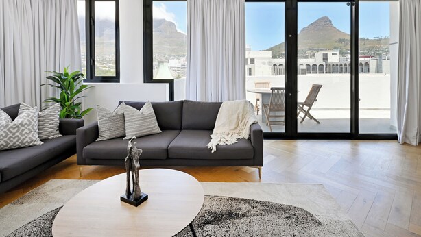 A modern living room with a grey sofa, a coffee table, and a view of a cityscape and mountains through large windows. - City Centre Rooftop Penthouse with 360° City & Mountain Views! (Cape Town)