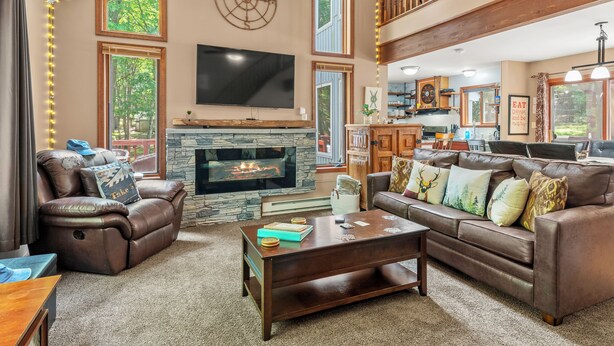 A spacious living room with a fireplace, leather sofas, a large flat-screen TV, and a kitchen area visible through an open door.