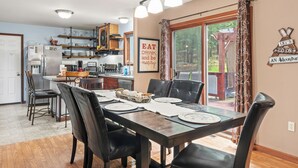 A dining area with a wooden table set for six, surrounded by black chairs. A kitchen is visible in the background with a refrigerator, stove, and shelves.