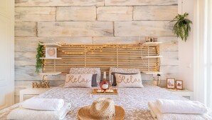 Una camera da letto con una testiera in legno, due cuscini con la scritta 'Relax', un comodino con una bottiglia e dei bicchieri, e un cappello di paglia sul letto.