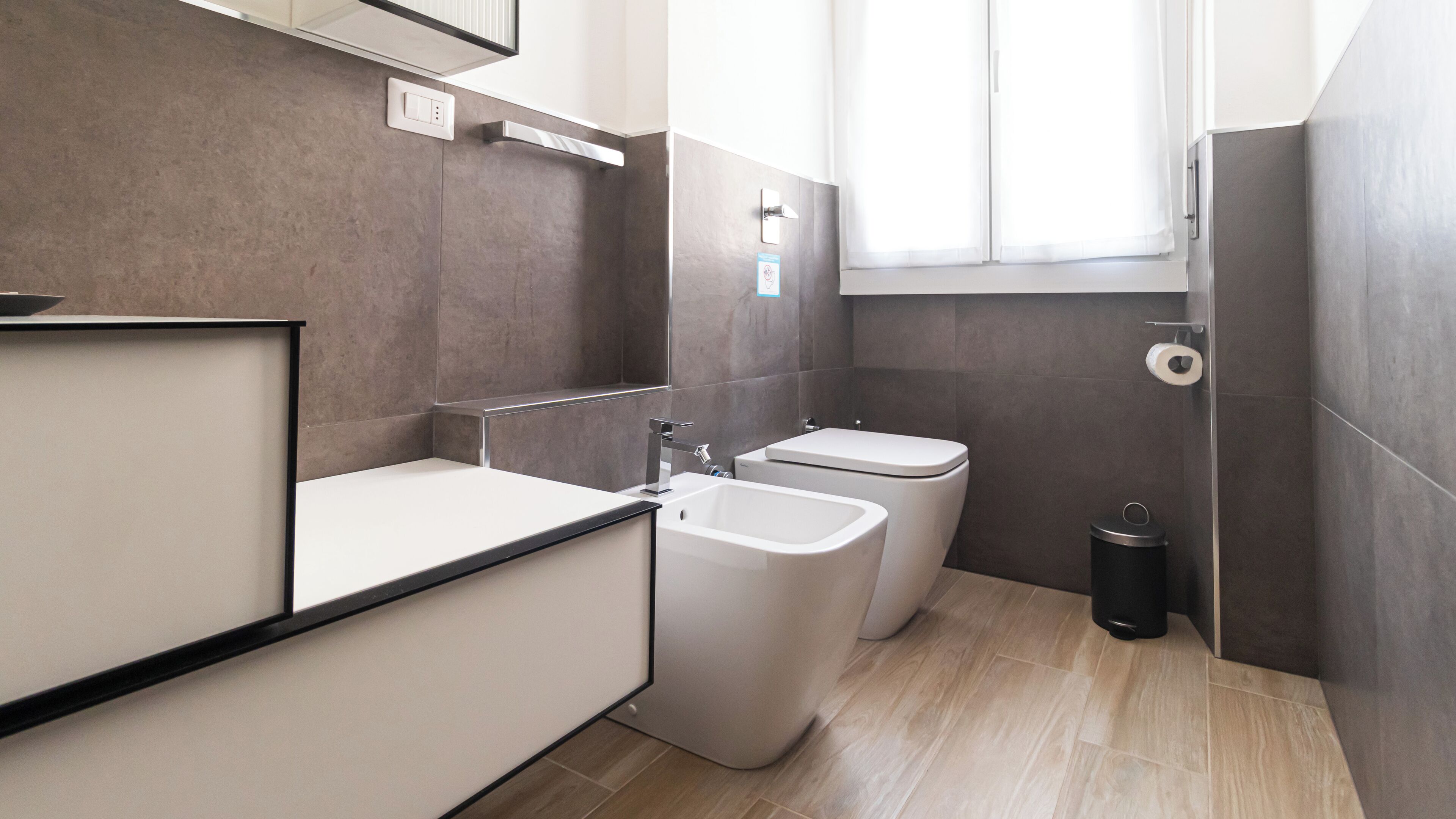 A modern bathroom with a bathtub, two toilets, a sink, and a trash bin.