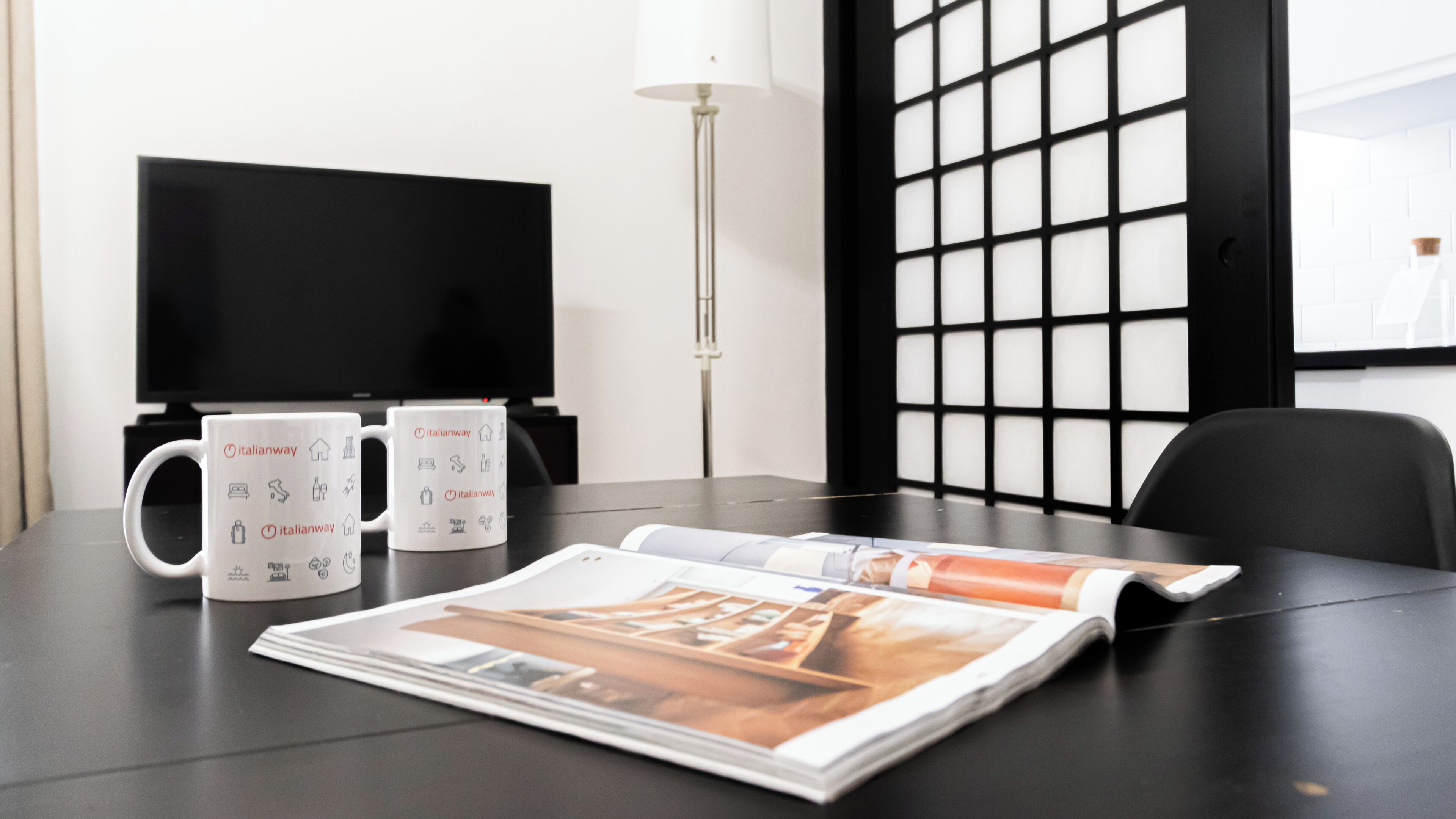 A hotel room with a black table, two white mugs with Italianway logos, a magazine, a TV, and a black chair.