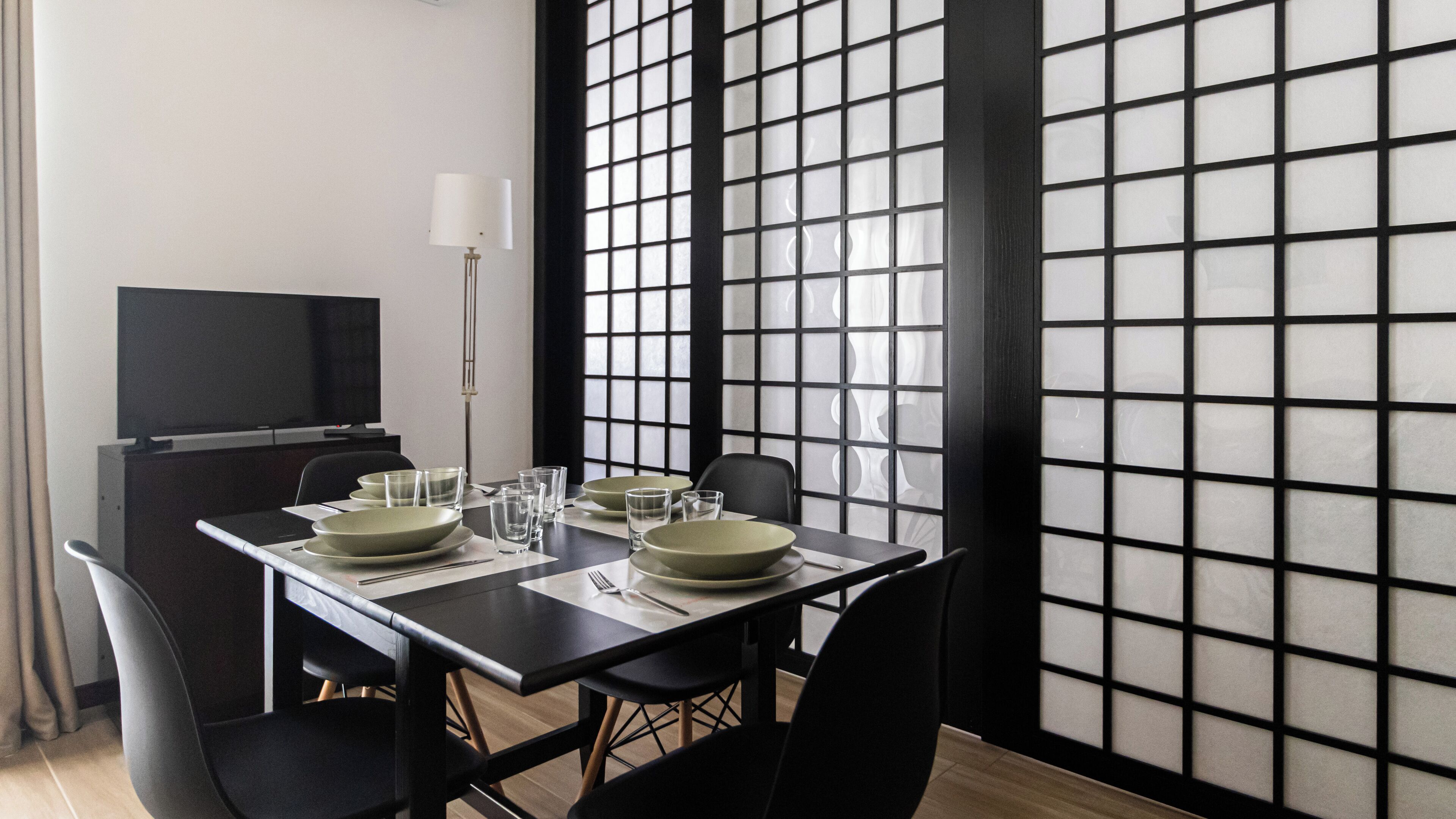Una zona de dinar moderna amb una taula negra preparada per a quatre, incloent plats, cuplets, vasos i coberts. Un televisor de pantalla plana està instal·lat a la paret a l'esquerra.