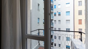 Une vue depuis un balcon doté d’une balustrade en métal, donnant sur un immeuble à plusieurs étages avec de nombreuses fenêtres, dont certaines sont équipées de volets.