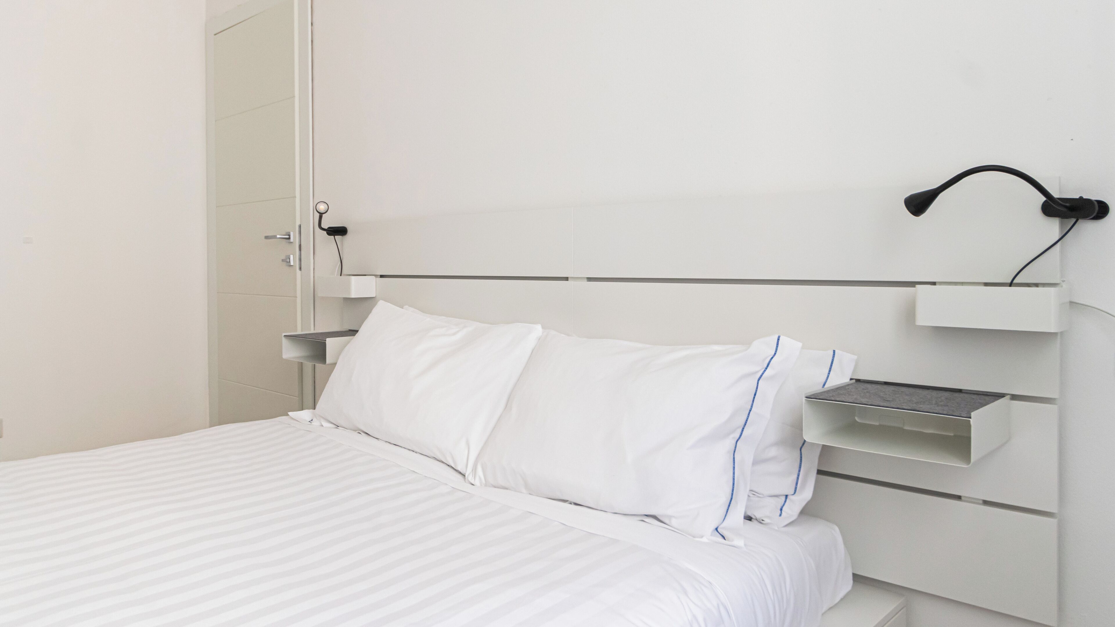 Una cama bien hecha con ropa blanca y cabecera con estantes integrados.